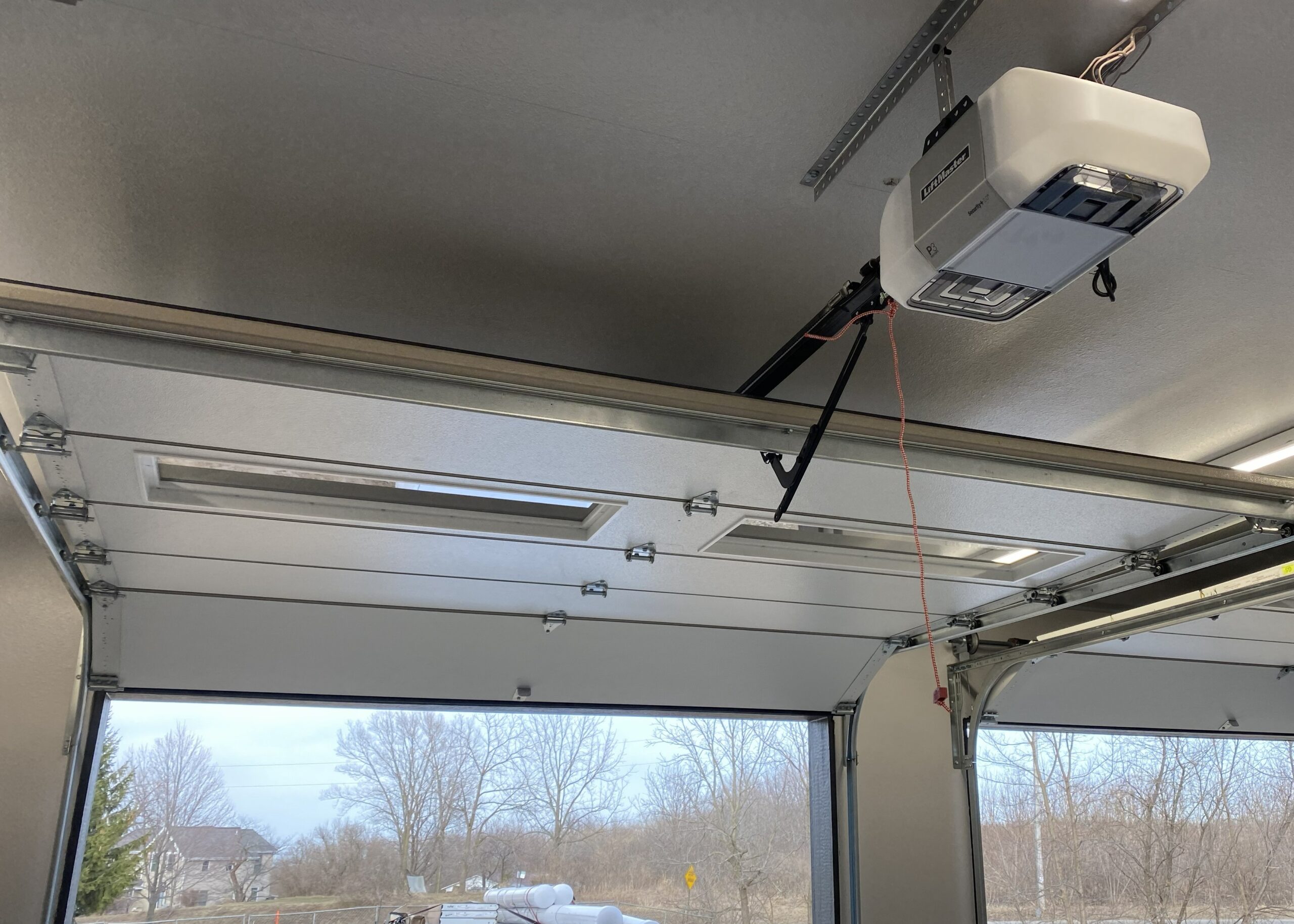 Interior of a garage with open overhead doors, a ceiling-mounted door opener, fluorescent lights, and a quiet winter landscape beyond.