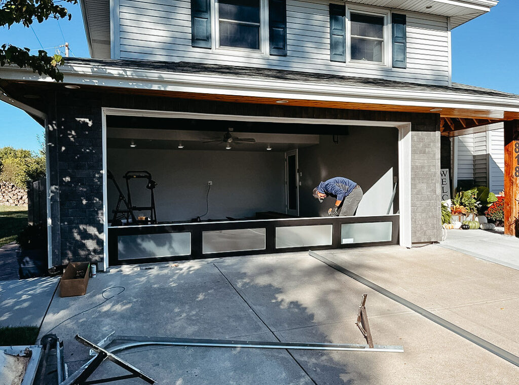 A worker completes a custom garage door installation on a modern home, with tools and materials scattered across the driveway in bright daylight.